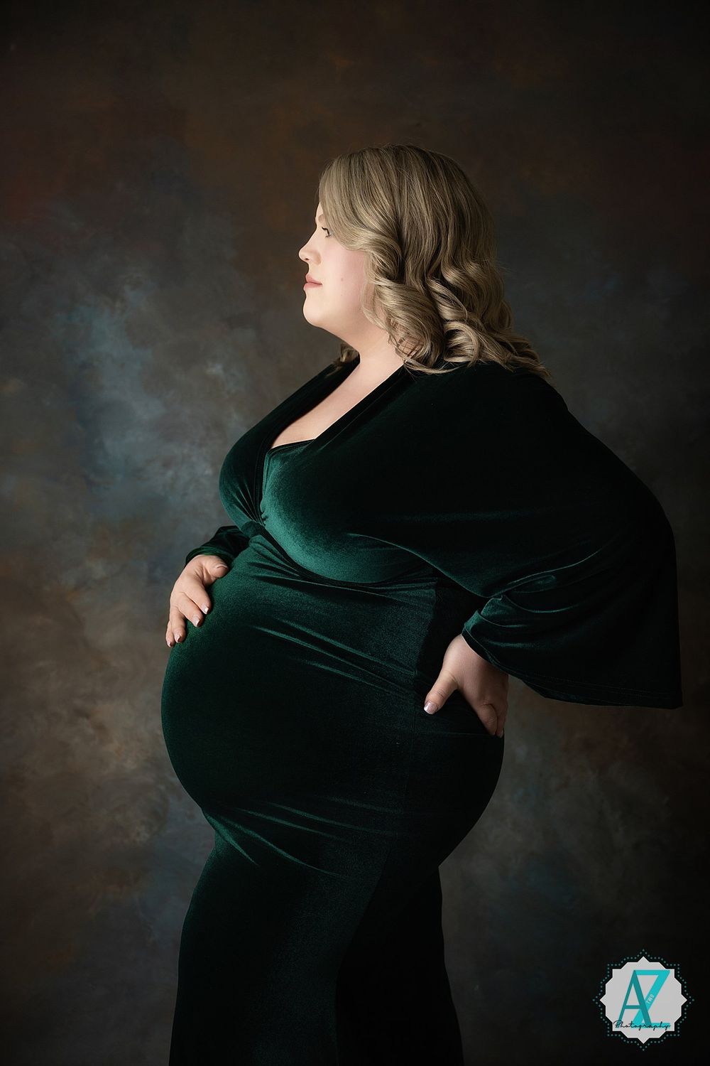 A pregnant woman poses in an elegant emerald velvet dress against a moody dark background in a maternity portrait.