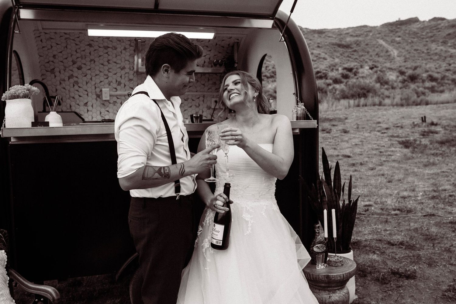 A couple in wedding attire sharing drinks by the back of an open vehicle in a desert landscape, captured in black and white.