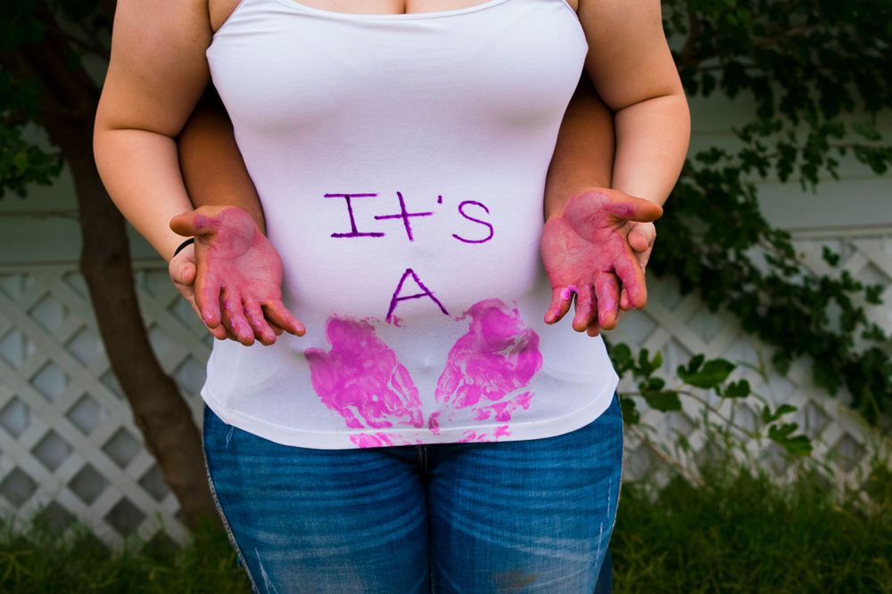 Expectant mother holding her stomach, with a white shirt featuring the phrase "It's a" for a joyful baby announcement.
