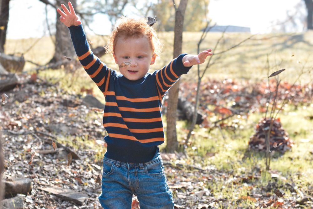 A red-haired boy stands in the woods, playfully tossing leaves into the air.