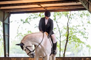 Equestrian rider in formal attire practicing with a white horse under a covered arena.