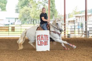 A barrel racer rides a white horse around a red and white stateline barrel in a dirt arena.