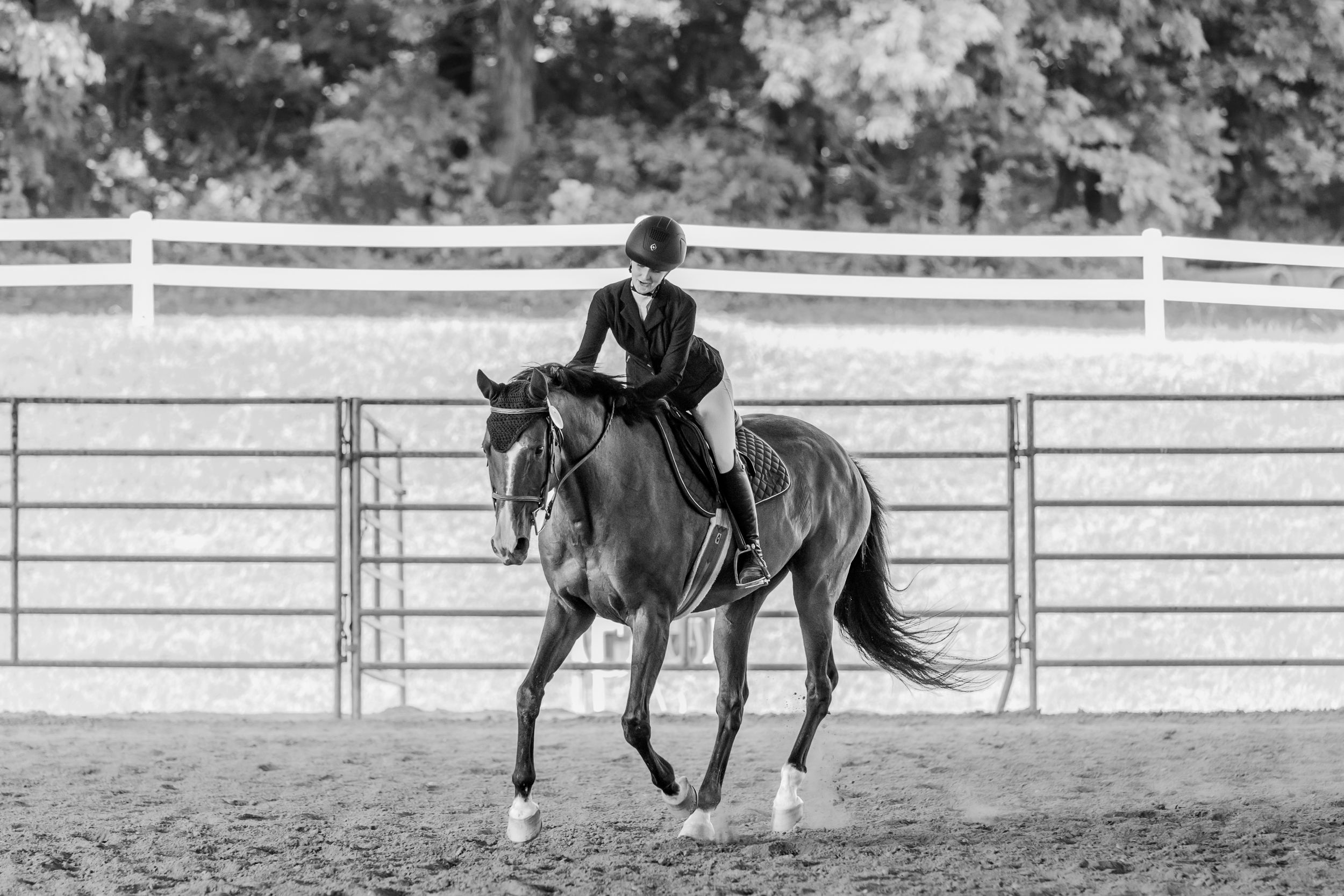 Black and white photo of an equestrian riding a horse in a ring with white fencing.