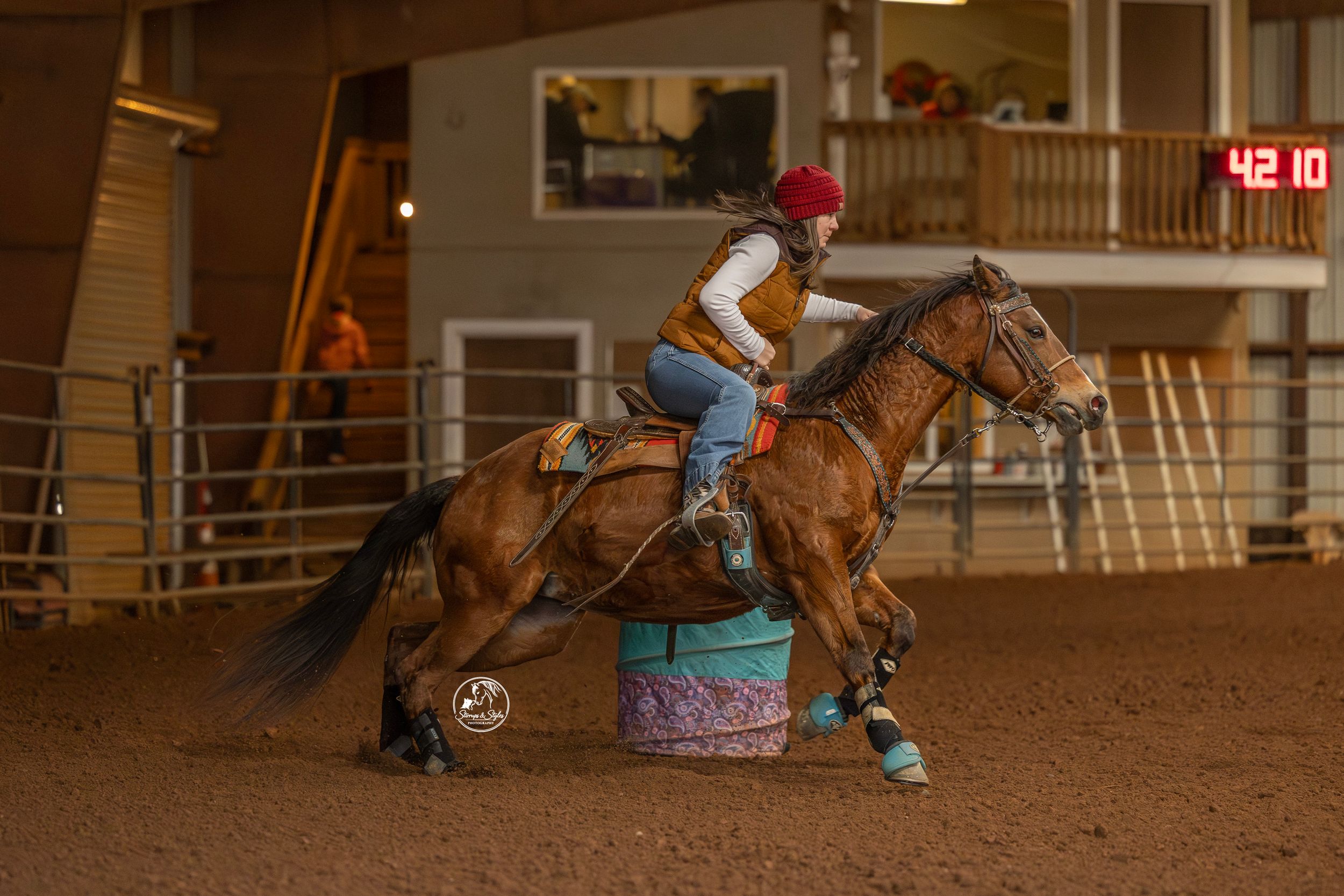 A rider on horseback competes in a barrel racing event inside a dirt arena with spectators watching from the stands.