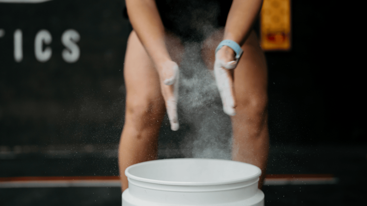 Hands applying chalk powder over a white bucket in a dark gym setting.
