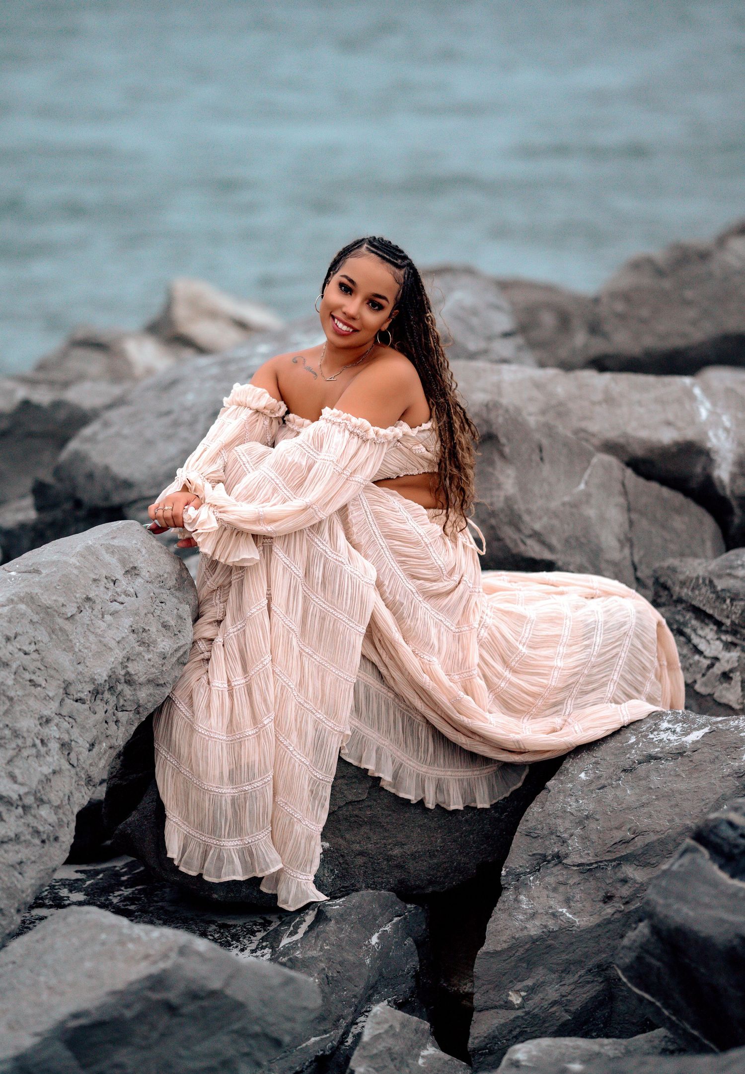 A feminine figure in a flowing off-shoulder cream dress poses gracefully on dark coastal rocks near the ocean.