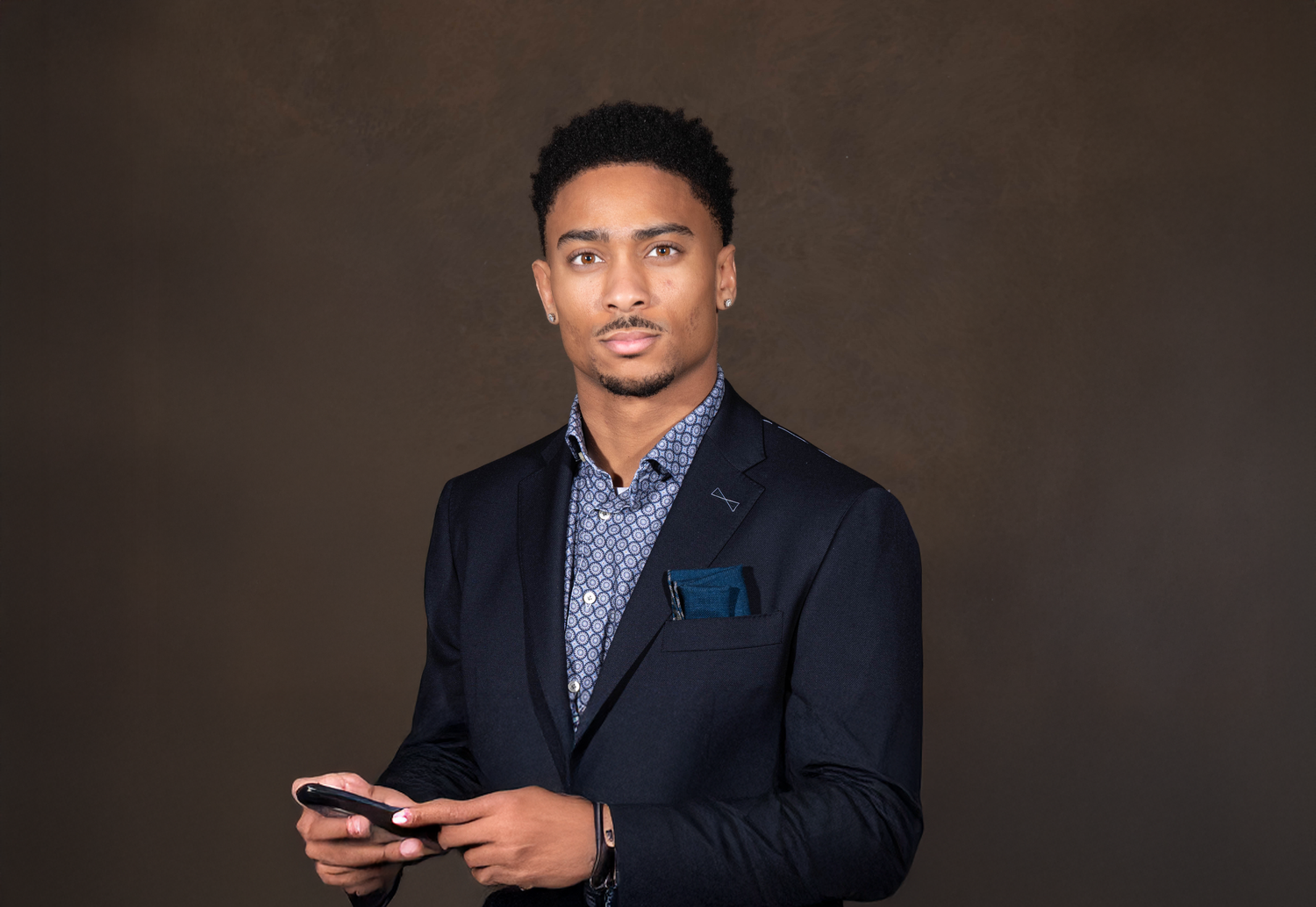 Professional wearing navy suit and patterned shirt holding phone against dark background.