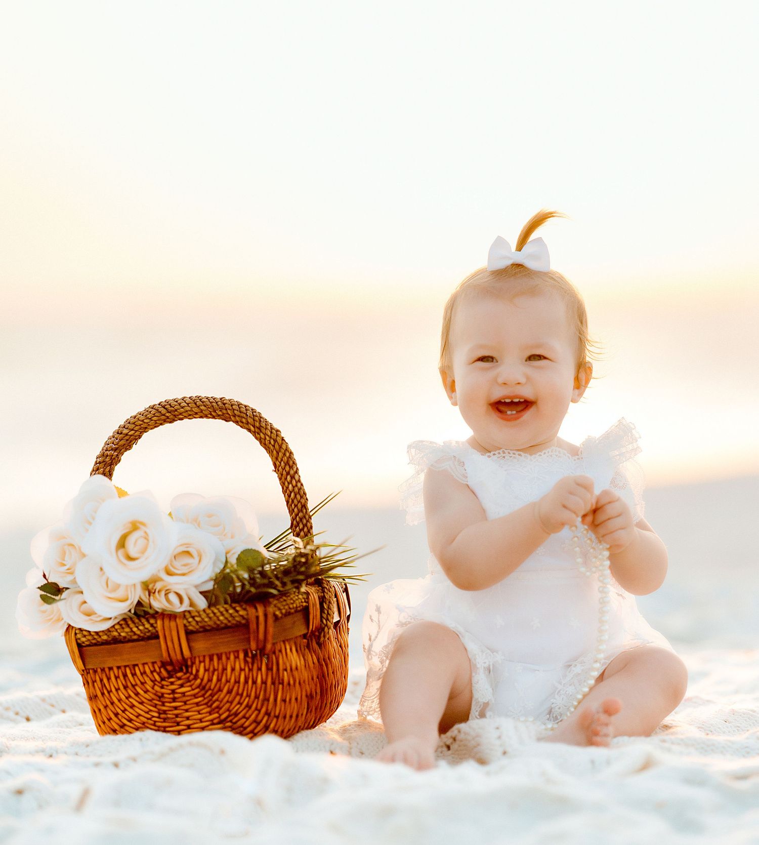 Baby in white dress sits on white sand beach next to wicker picnic basket during sunset.