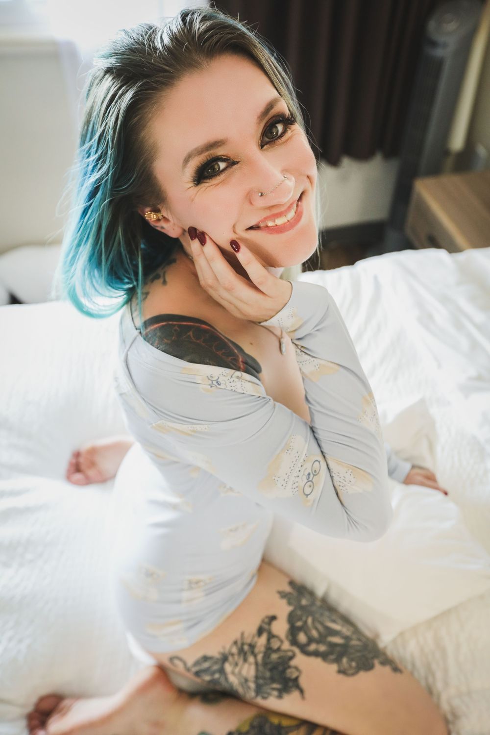 A person with blue-tipped hair wearing a white long sleeve top poses on white bedding, showing visible tattoo art.
