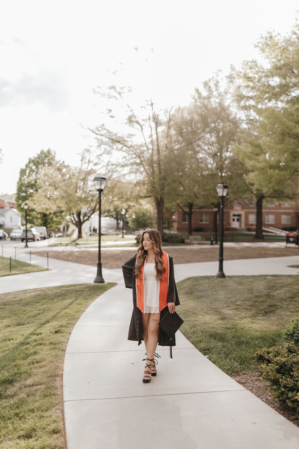 Graduate in academic regalia walks along curved campus sidewalk during graduation photoshoot. Virginia graduation/senior session.