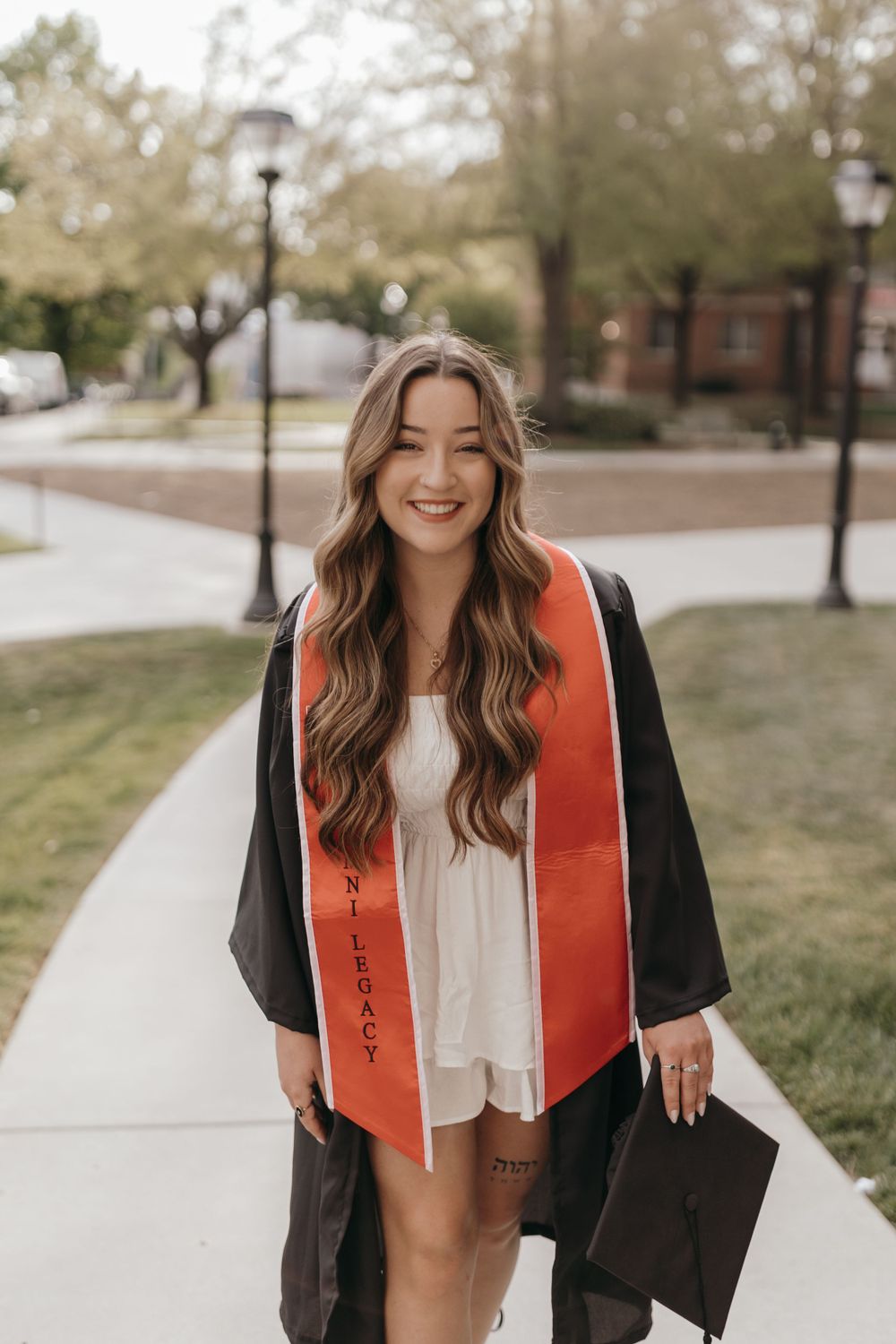 Graduate wearing cap and gown with orange graduation stole smiles on university campus pathway. Virginia graduation/senior session.