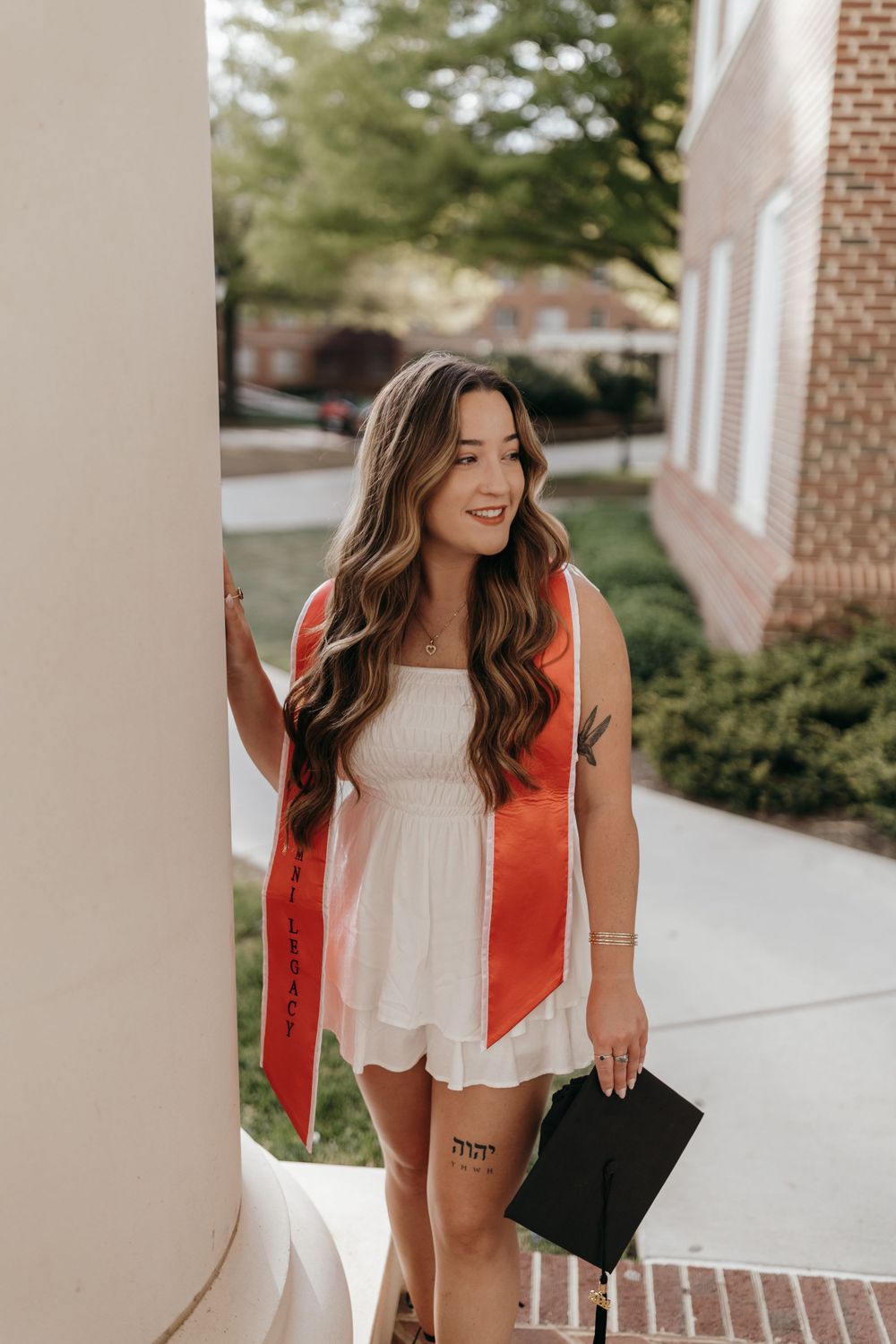 Graduate in white dress and orange stole poses between classical columns during graduation photoshoot. Virginia graduation/senior session.