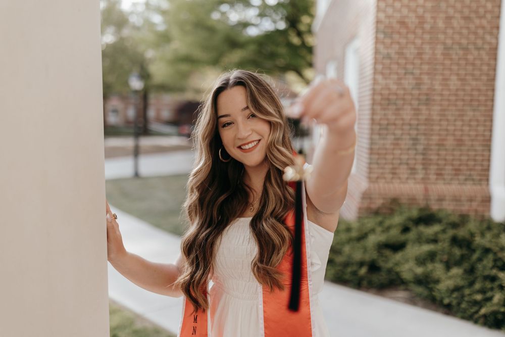 Graduate with long wavy hair smiles joyfully while posing between white columns. Virginia graduation/senior session.
