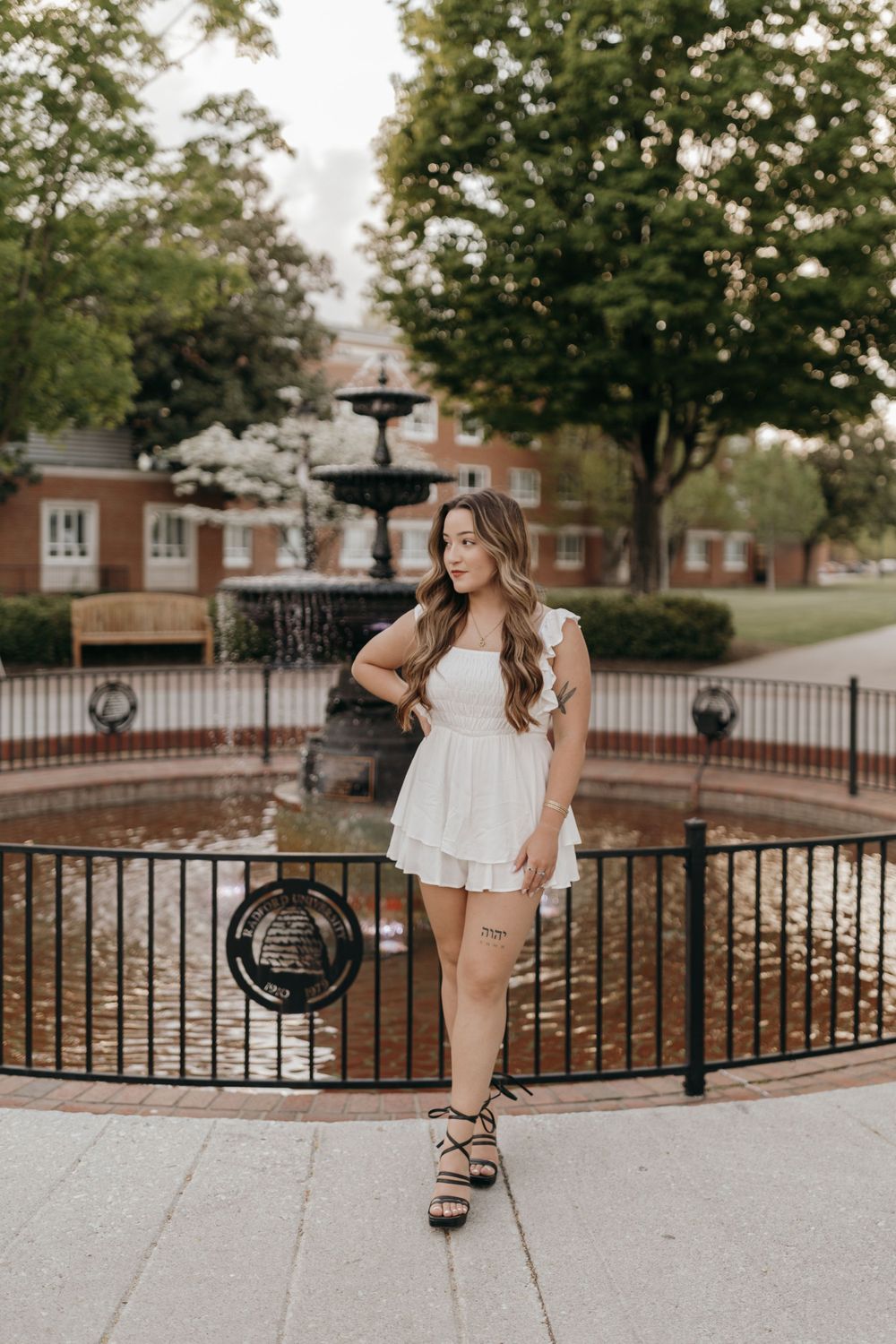 Graduate in white dress poses by ornate campus fountain during graduation photo session. Virginia graduation/senior session.