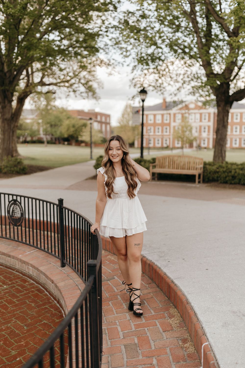 Graduate in white dress poses on brick pathway near wrought iron railing with red buildings in background. Virginia graduation/senior sessio
