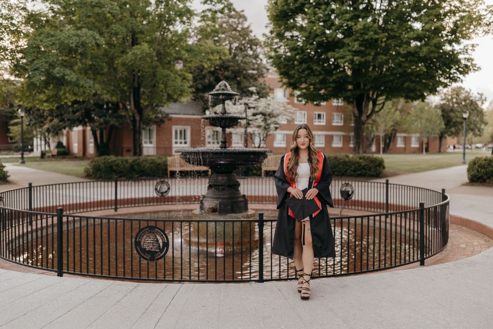Graduation photoshoot by ornate fountain with brick buildings and iron fence in background. Virginia graduation/senior session.