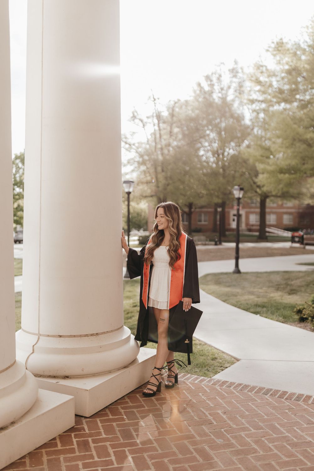 Graduate in orange stole and white dress poses by classical column on campus walkway. Virginia graduation/senior session.