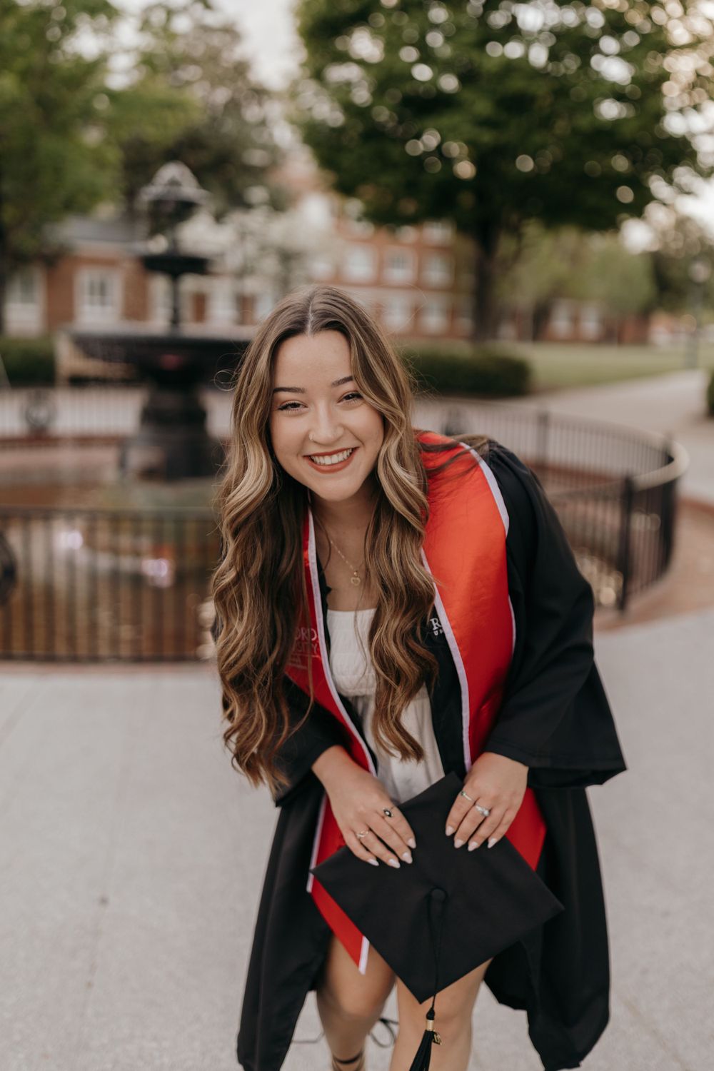 Graduate wearing graduation gown and displaying cap in front of campus fountain. Virginia graduation/senior session.
