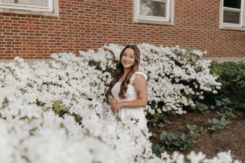 Graduate poses among blooming white flowers near brick university building. Virginia graduation/senior session.