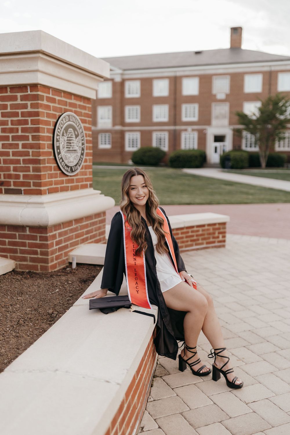 Graduate in academic regalia sits on brick wall by university seal. Virginia graduation/senior session.