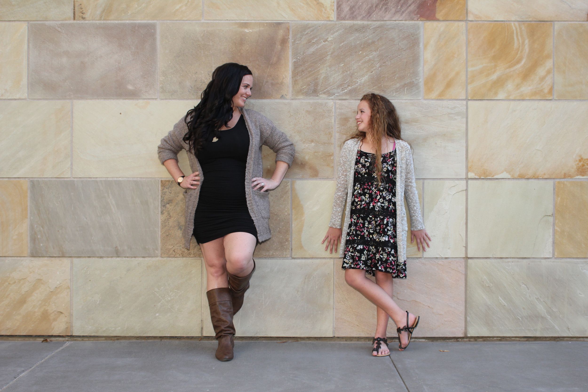 Mom and daughter in stylish dresses and boots lean against a stone wall during a casual photo shoot. Family portrait.
