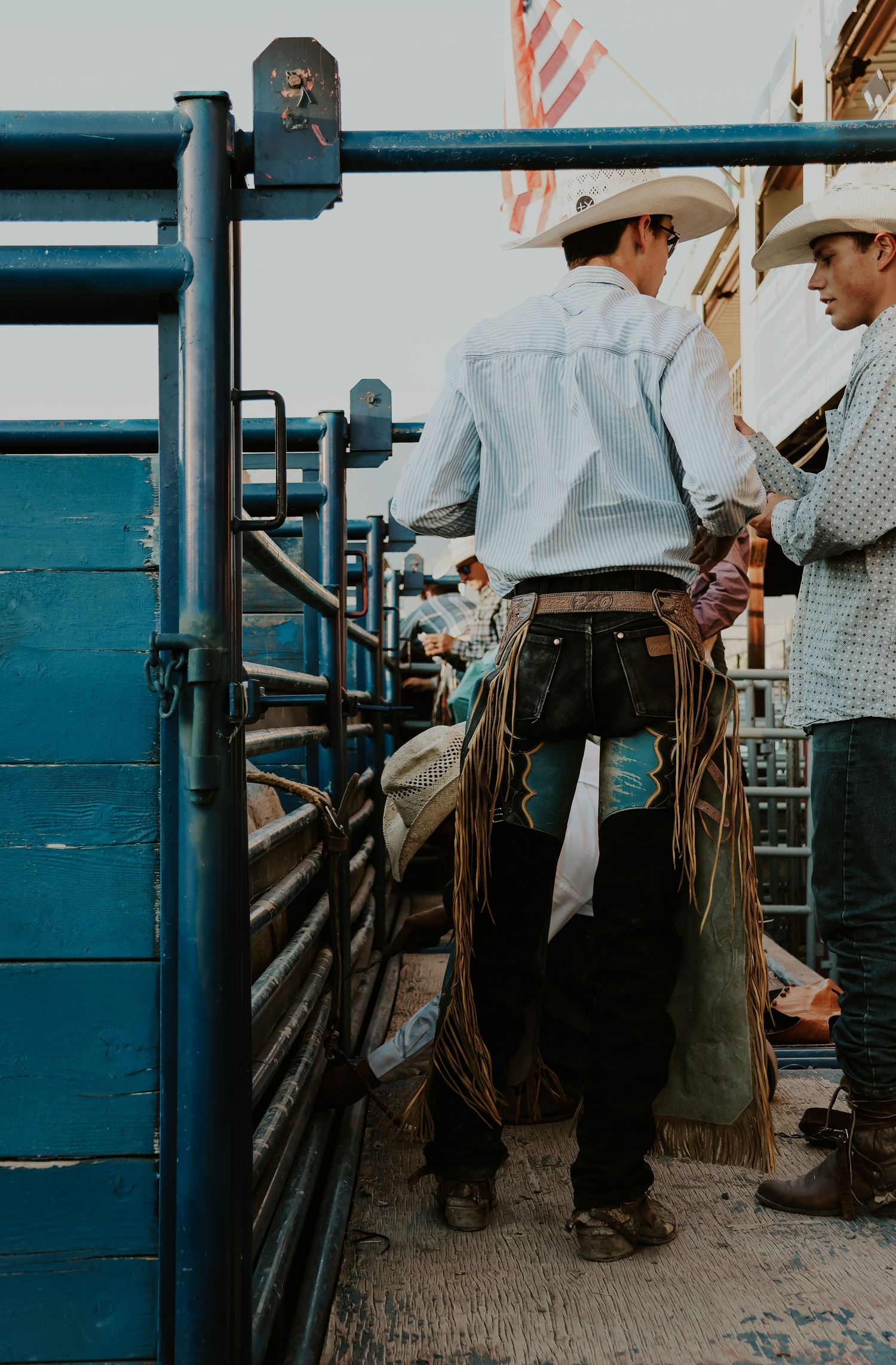 Cowboys in western attire stand near blue metal gates at a rodeo.