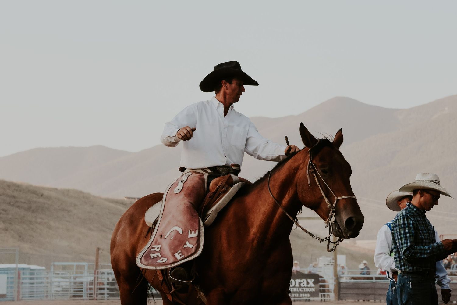 Rider mounted on brown horse at a dusty ranch arena.