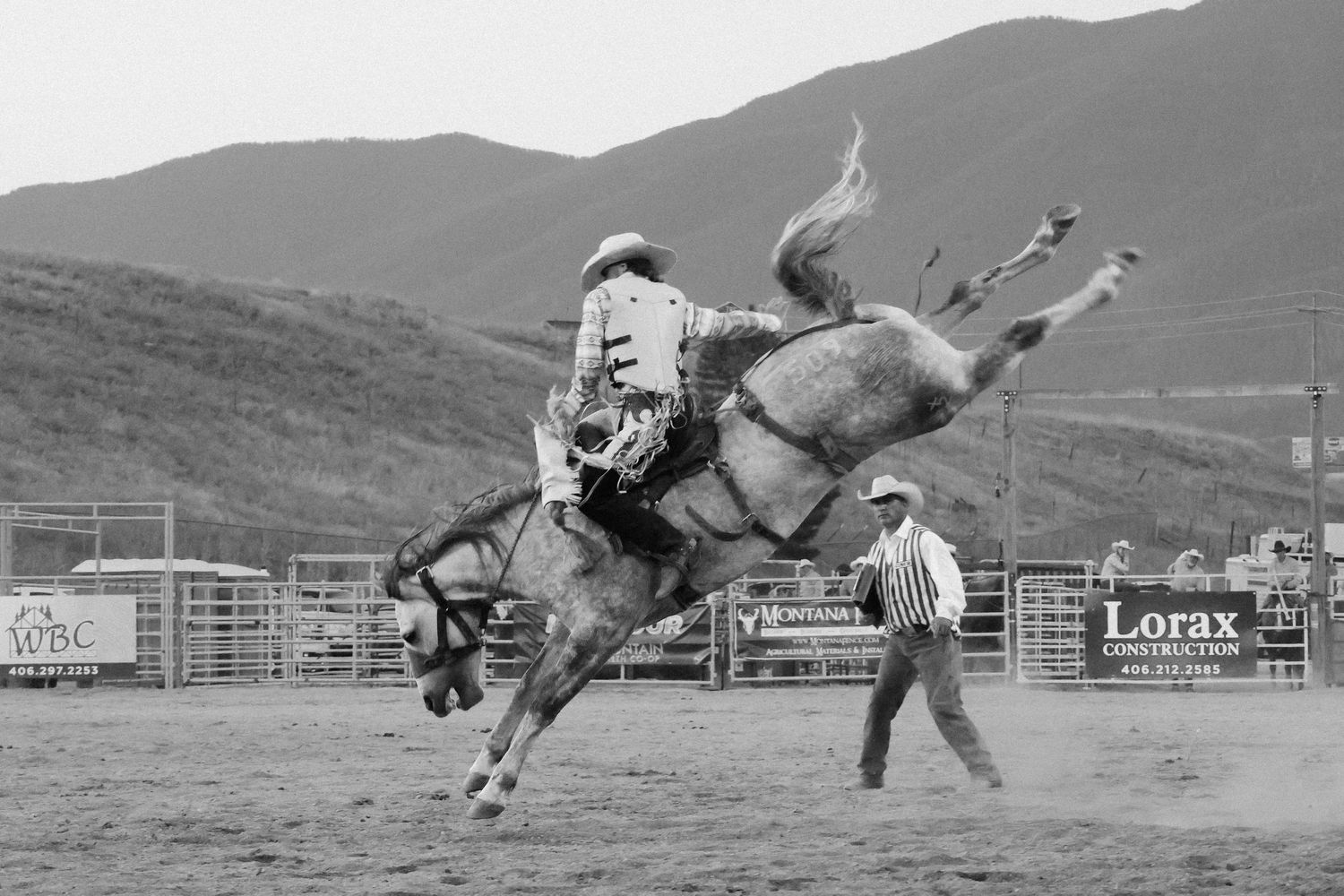 Black and white photo of rodeo rider on bucking bronco with mountains in background.