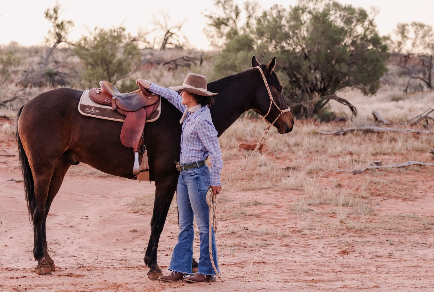 People in western attire stand with a brown horse in a desert landscape.