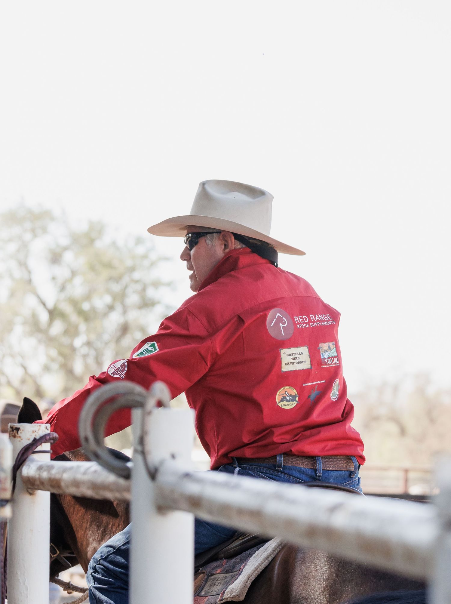 Cowboy in red jacket and hat sitting on horse by arena fence.
