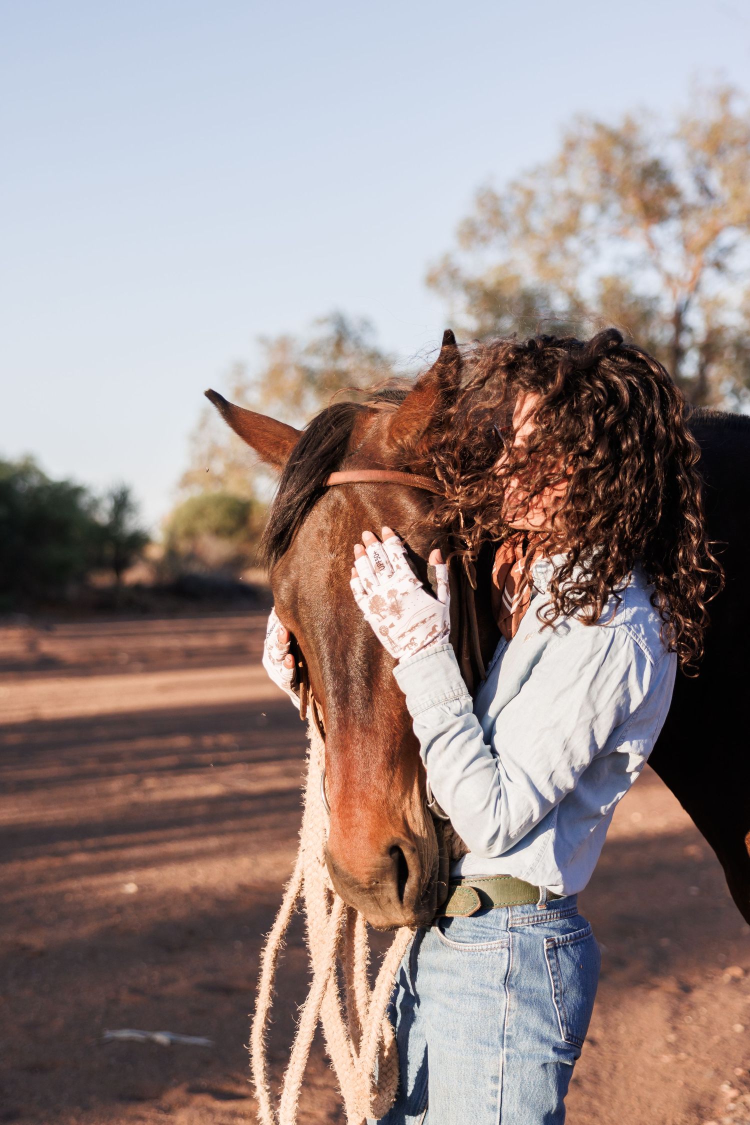 Person in denim and white shirt affectionately petting a brown horse outdoors.