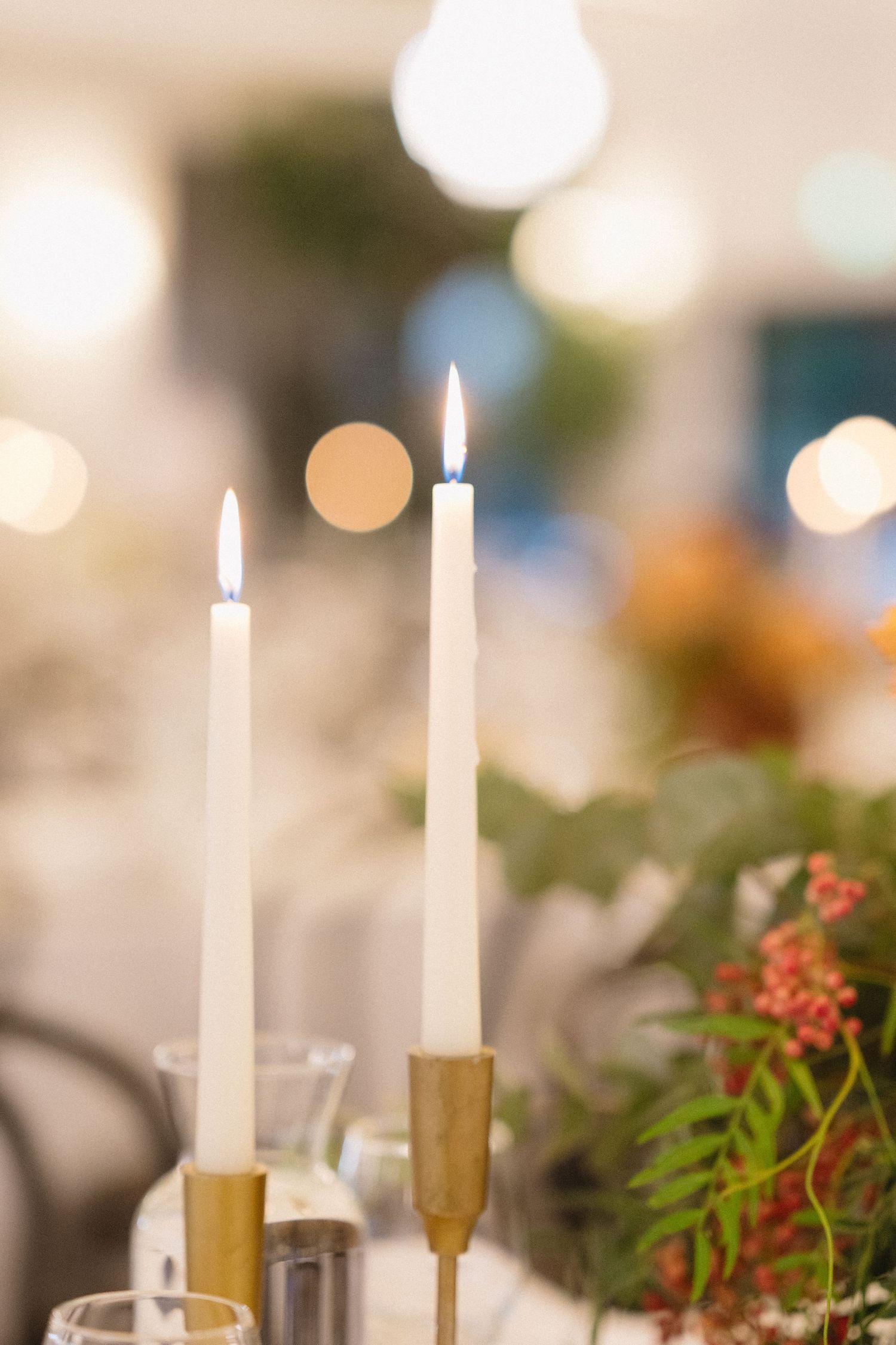 Two lit white taper candles in gold holders create an elegant tablescape with bokeh lights in background.