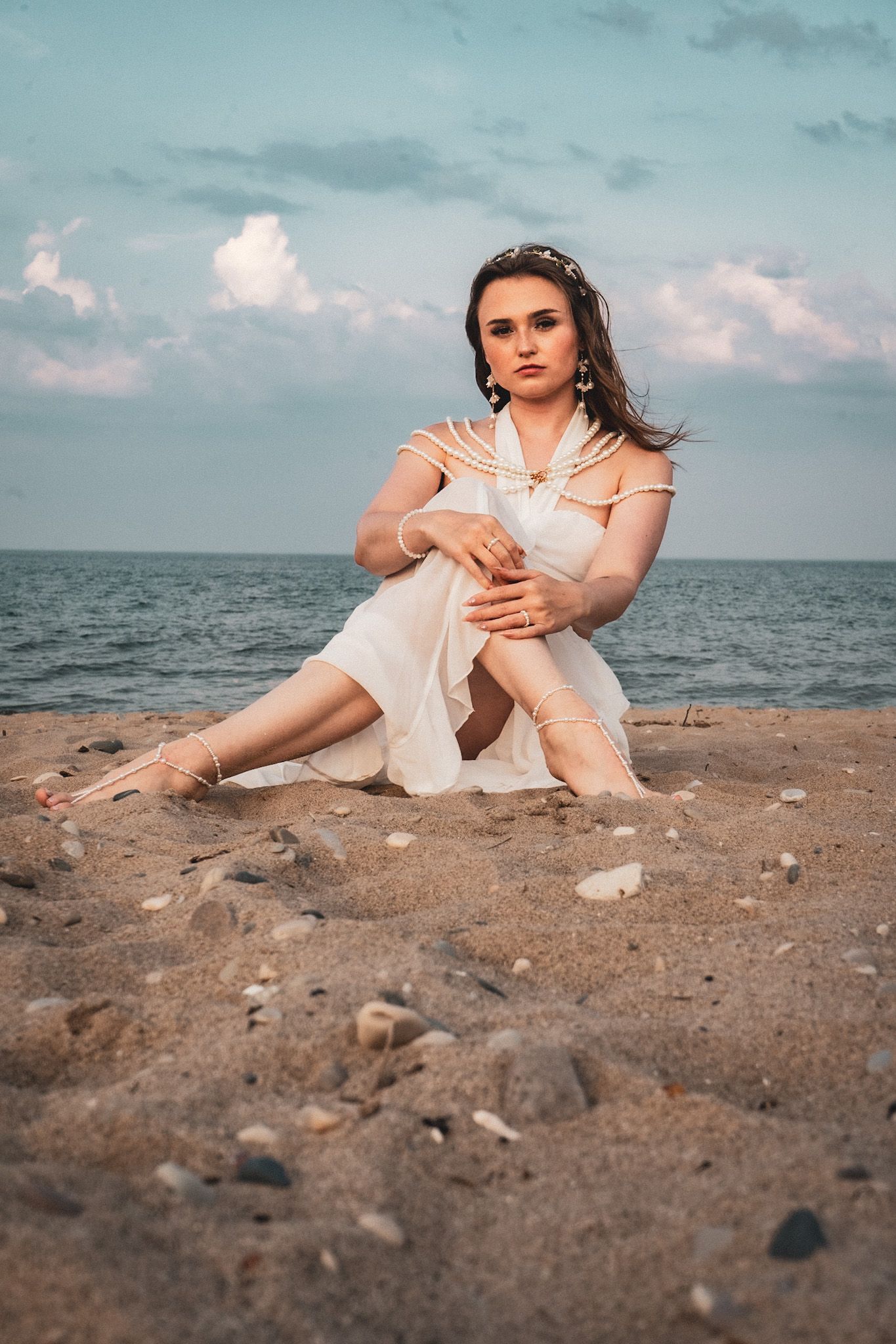 Person in white dress sits contemplatively on sandy beach against ocean backdrop.