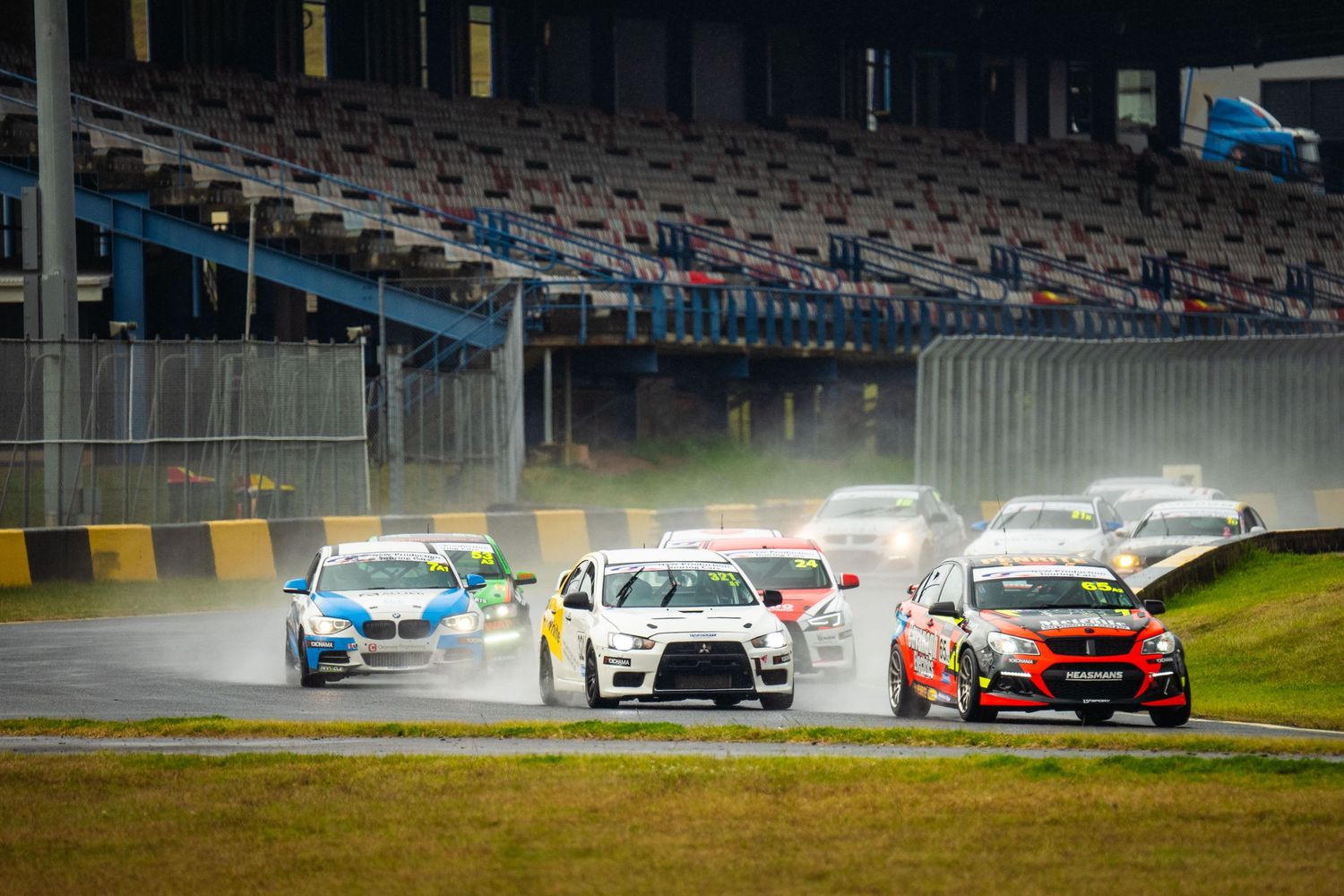 Race cars compete in wet conditions on a racing circuit with spectator stands visible in the background.