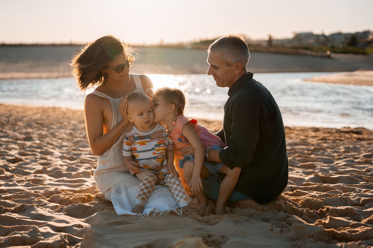 girl kisses her toddler brother while they are sitting at parents laps with a beautiful sunset at north curl curl