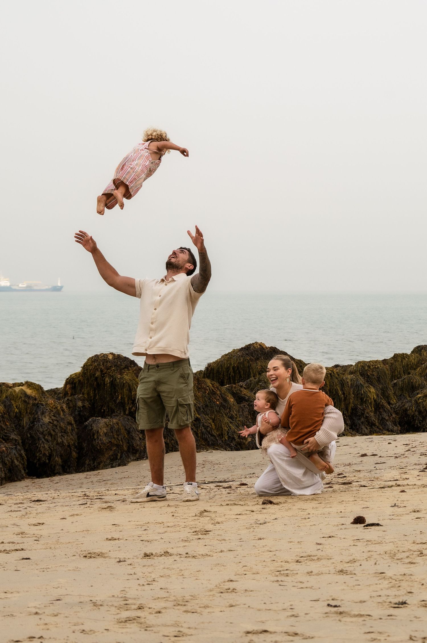 Playful family moment as baby is tossed in the air at beach with rocks and ocean backdrop.