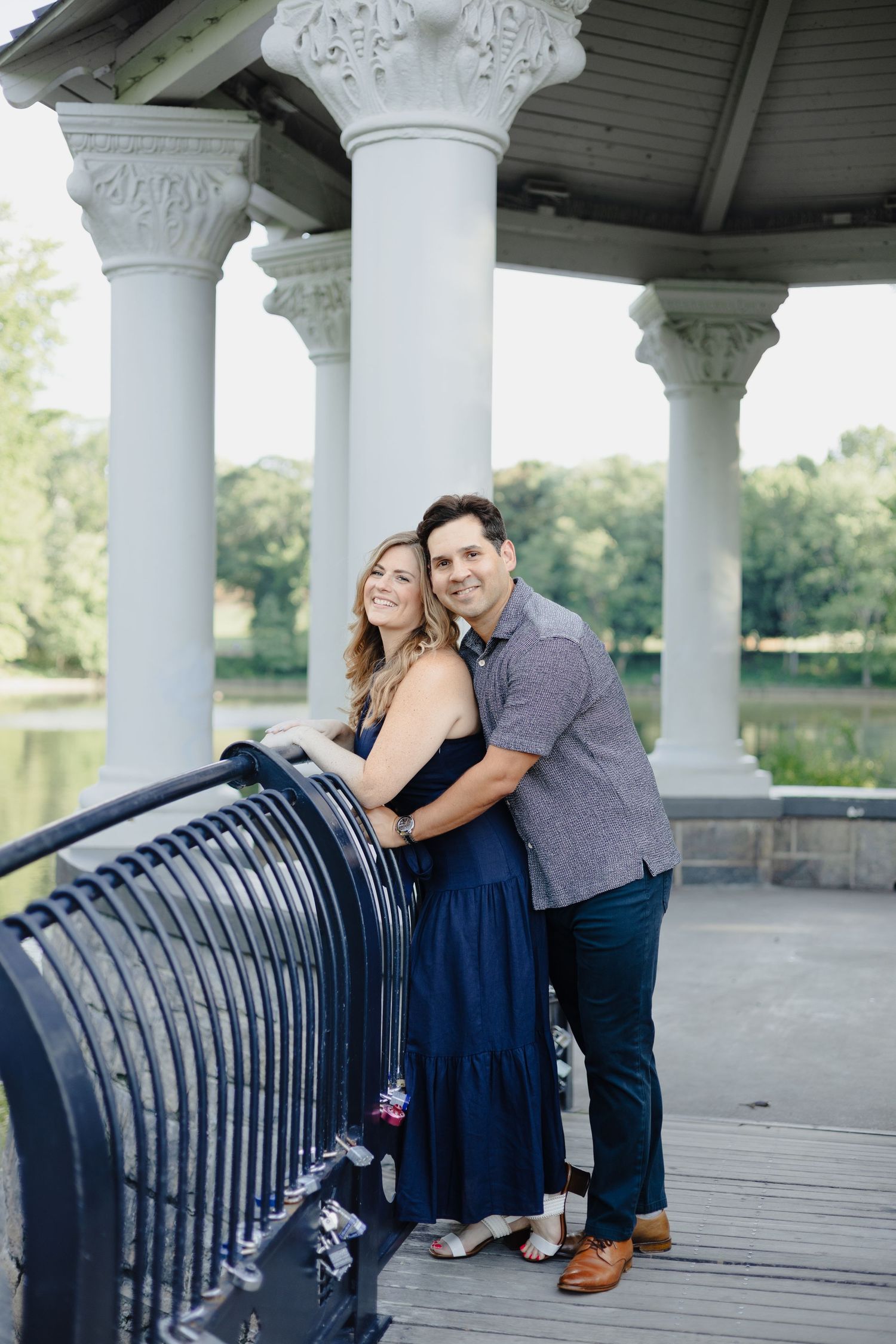 A couple stands together in a sunny meadow surrounded by green trees.