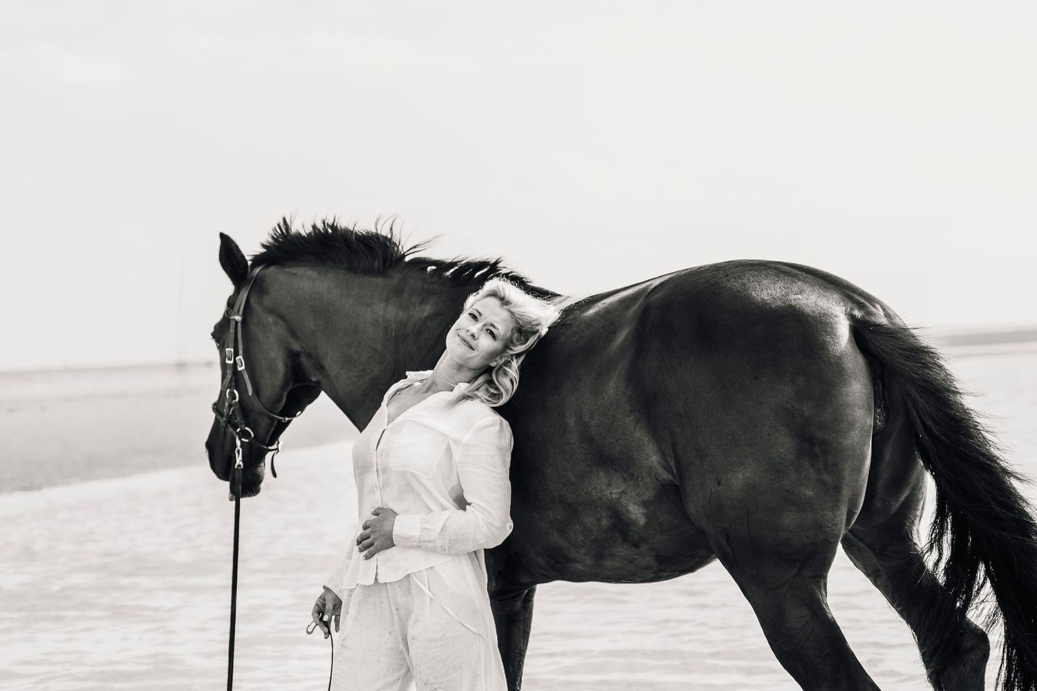 A black horse and person in white pose together on a sandy beach in monochrome.