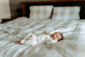 A small baby wearing white sleeps peacefully on white bedding in a cozy bedroom. Lifestyle session.
