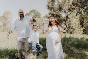 A family playing and running together outdoors in a sunny field, with a woman in a white dress. Maternity session in Traralgon.