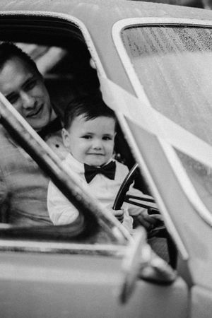 Black and white photograph showing a toddler in a bowtie sitting in a vintage car's front seat with an adult. Wedding at Valley View Manor.