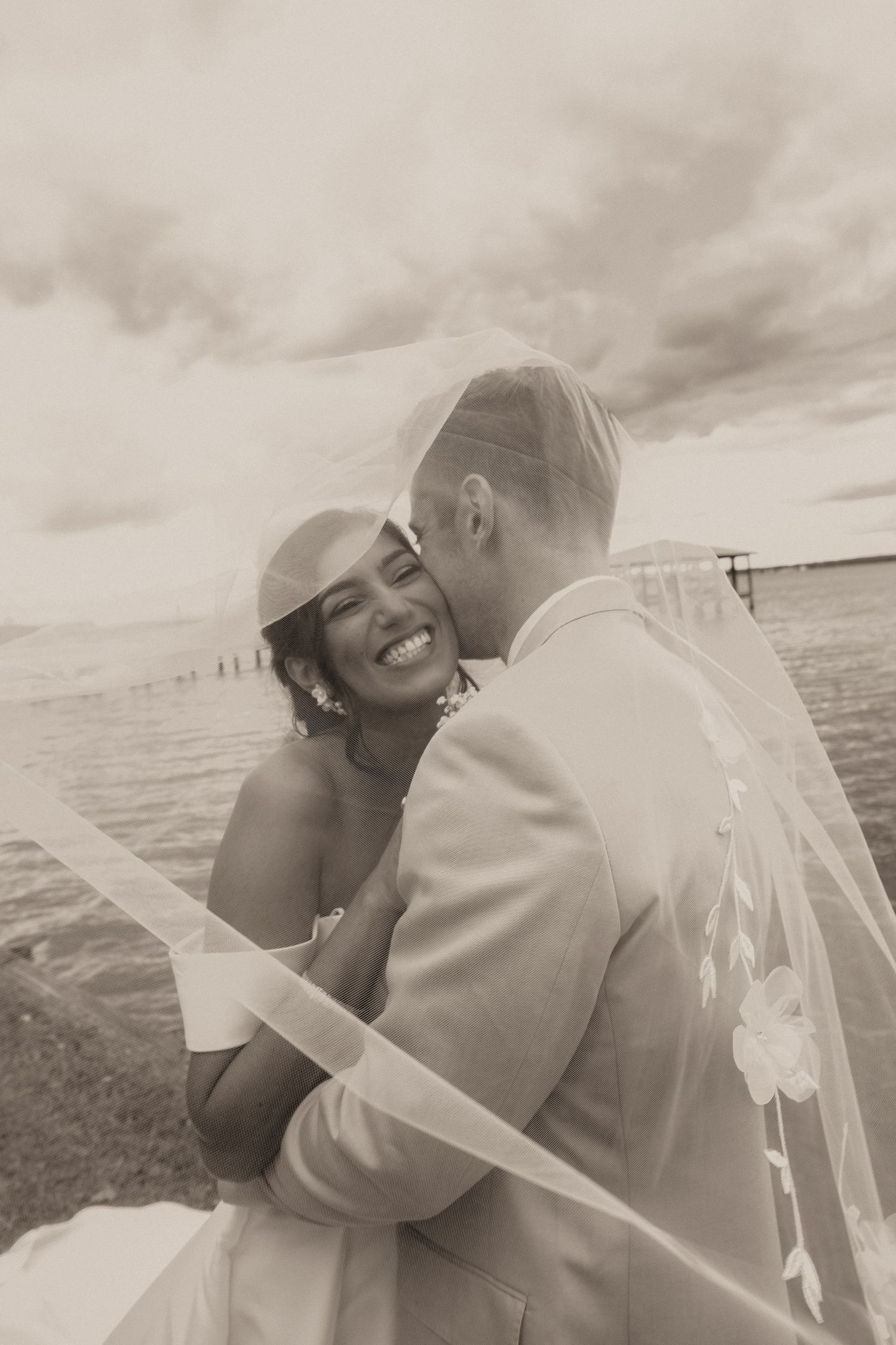 Black and white photo of groom kissing smiling bride near waterfront under cloudy sky.