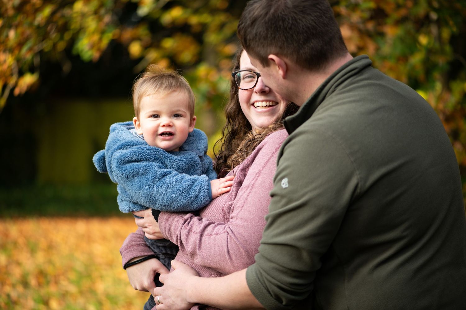 Mum holding 10 month old son, whilst looking lovingly at her husband during an Autumn shoot with rich autumn colours