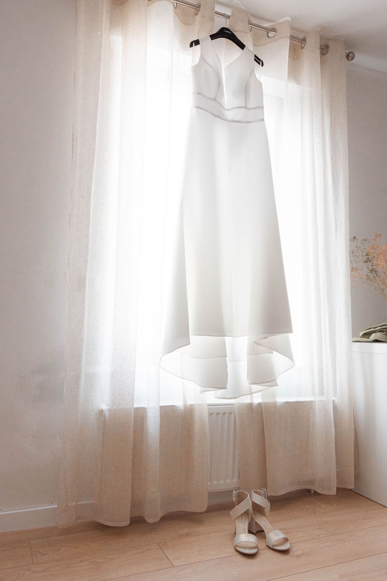 A pristine white wedding dress hangs by a window with bridal shoes placed beneath.