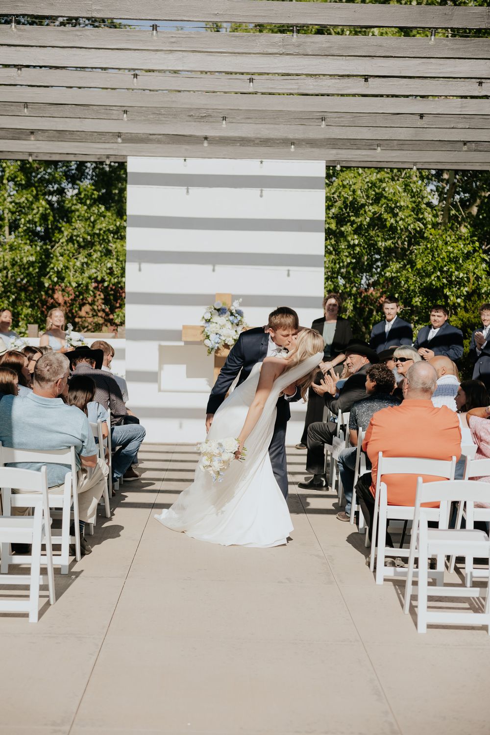 Bride and groom share a romantic dip kiss at outdoor wedding ceremony aisle with white chairs and floral backdrop.