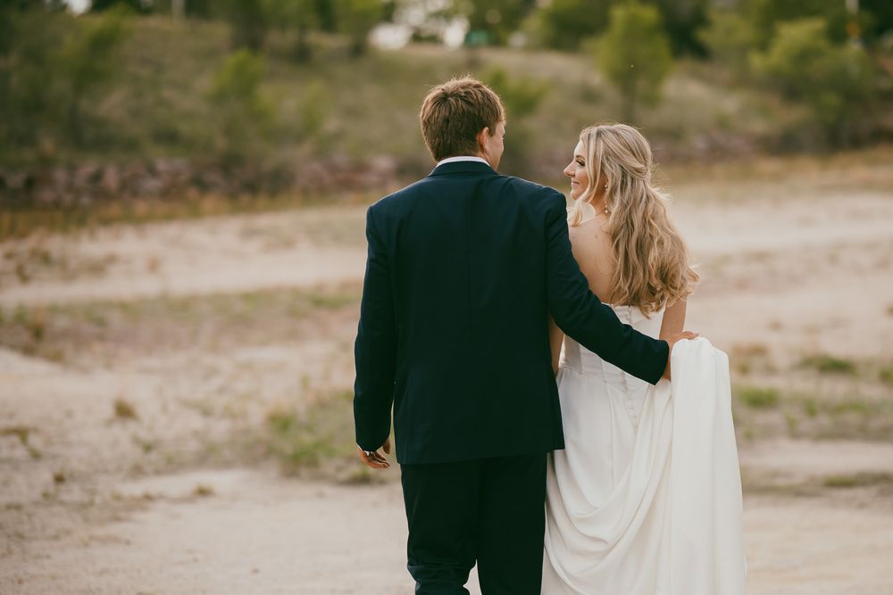 A bride and groom walking hand in hand on a sandy path, viewed from behind, dressed in wedding attire.