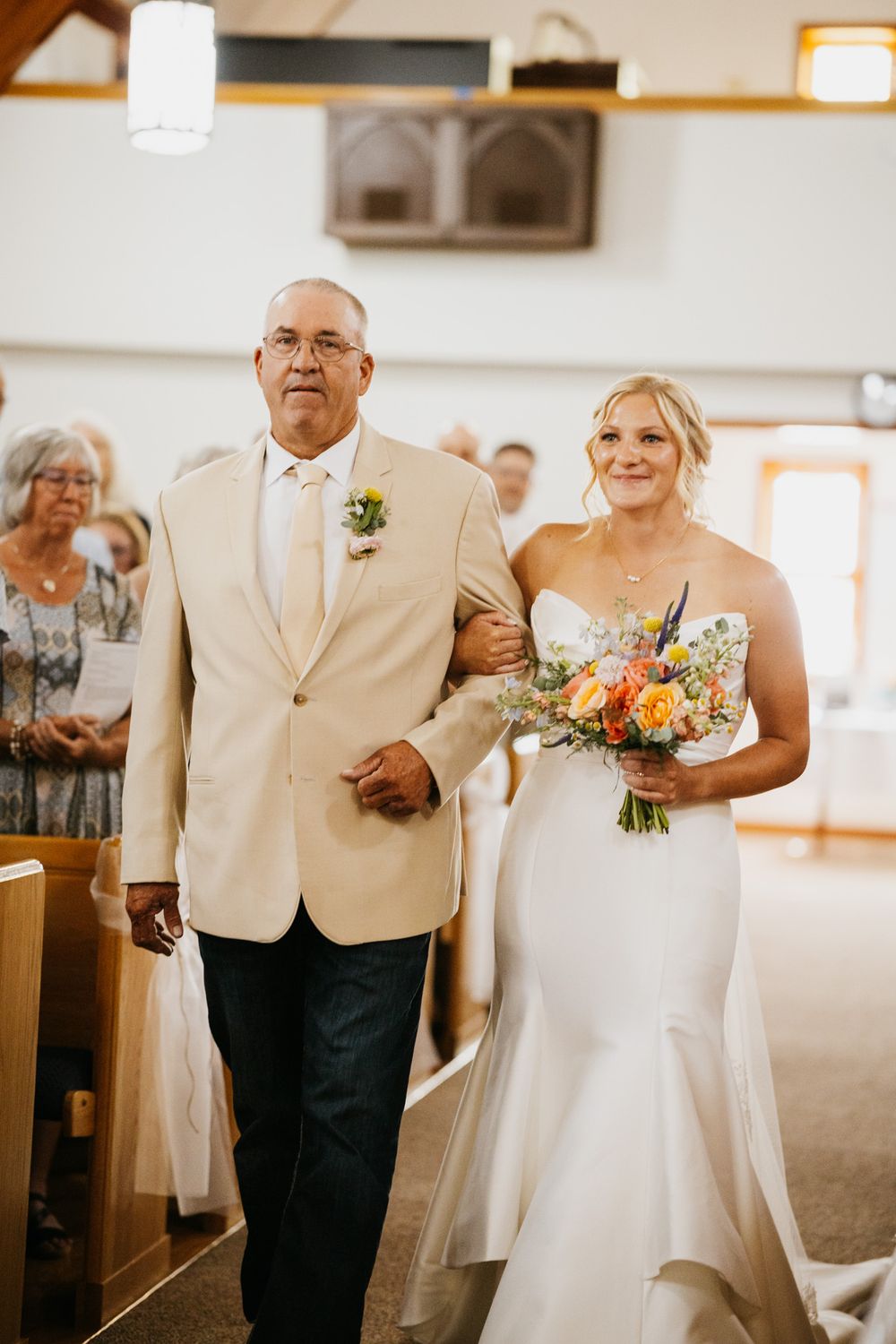 Bride in white gown holding colorful bouquet walks down church aisle with escort in cream blazer.
