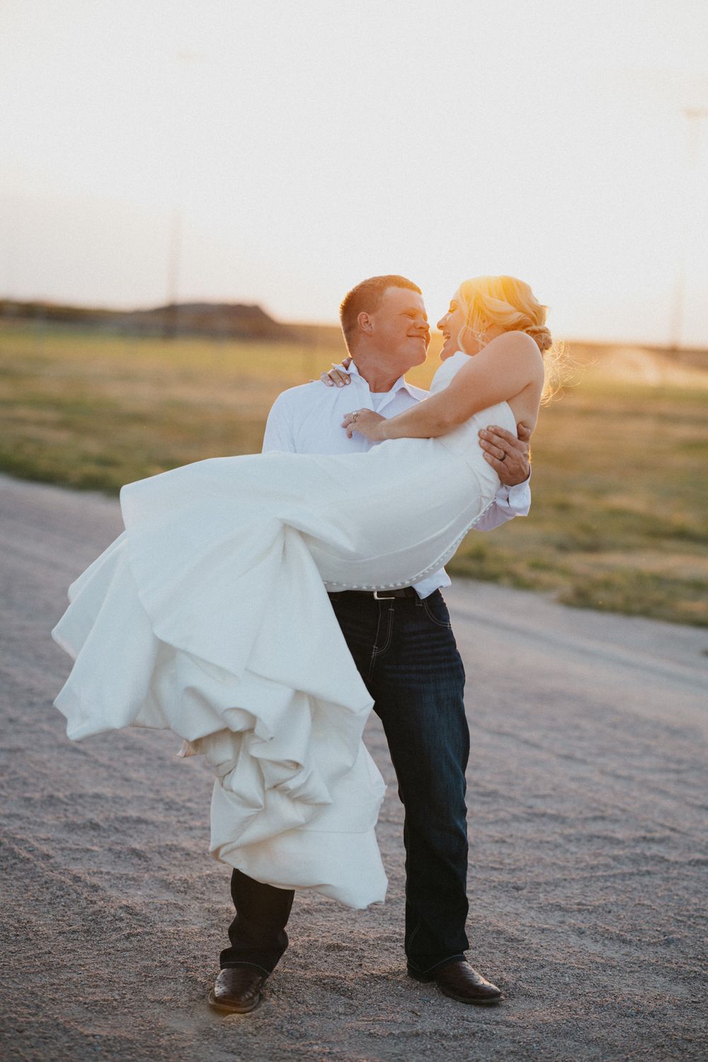 Groom lifts bride in white wedding dress at sunset on a gravel road, sharing a romantic kiss outdoors.