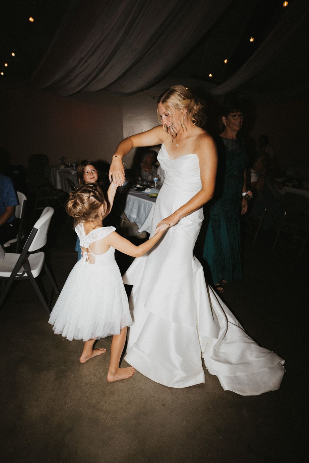 Bride in white gown dancing with young flower girl in white dress at wedding reception.