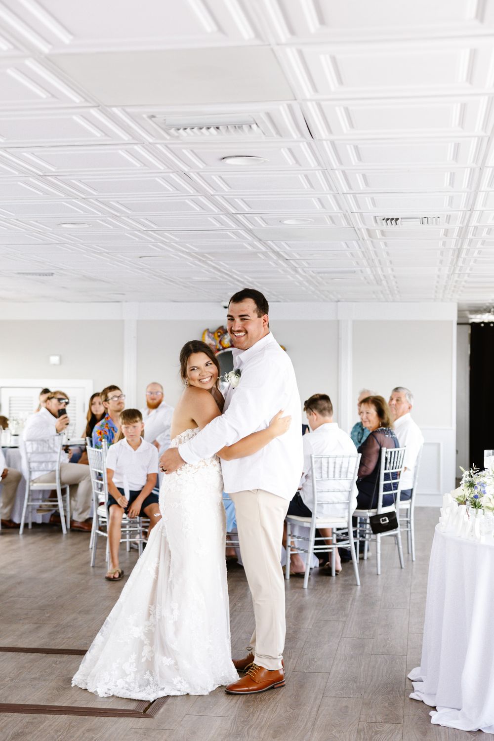 Bride and groom share first dance at elegant white wedding reception venue with guests seated at tables.